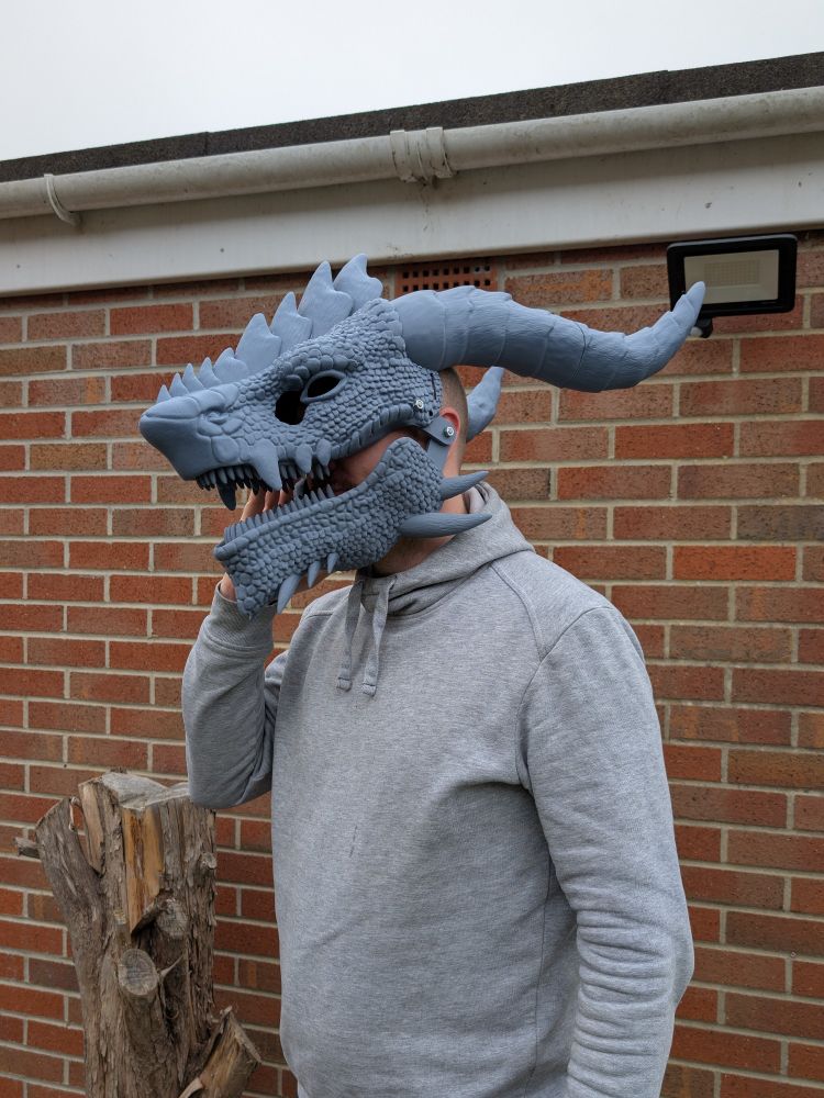 A person in a grey hoodie wearing a grey 3D printed dragon head mask. It has 2 large horns, plates along the nose, and spikes on the jaw and chin. The mouth is open to show sharp teeth. 