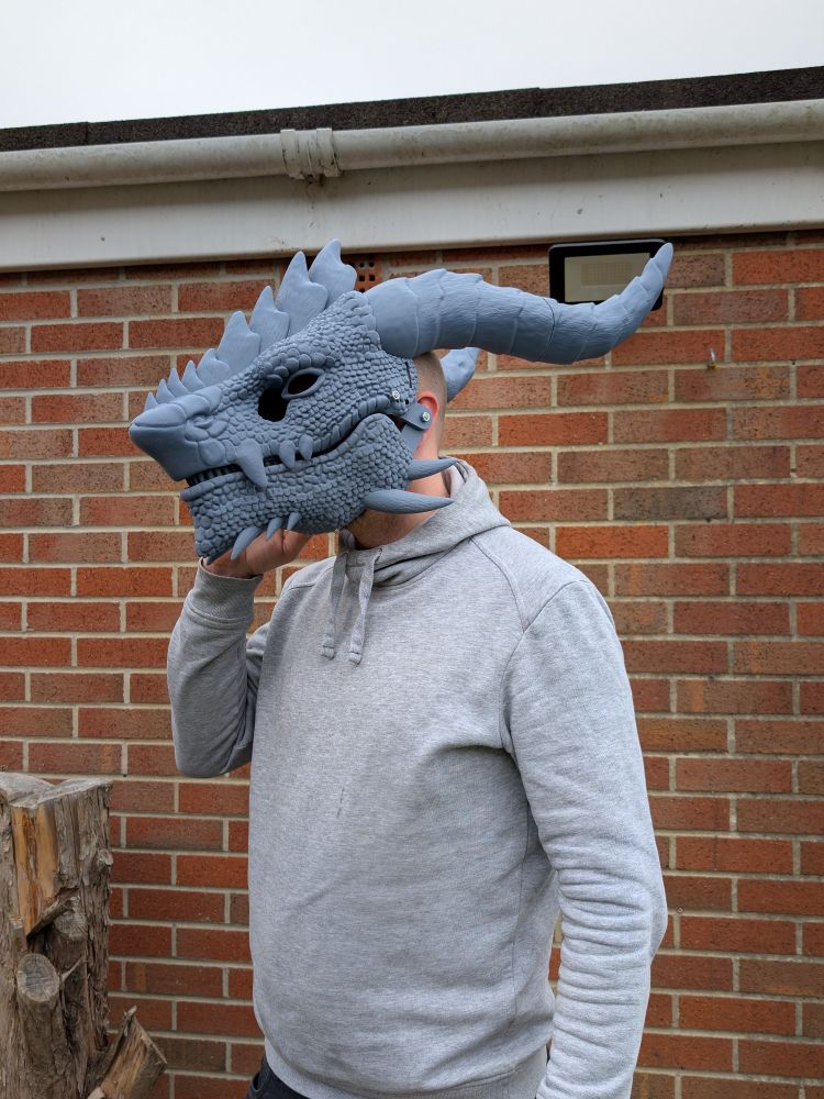 A person in a grey hoodie wearing a grey 3D printed dragon head mask. It has 2 large horns, plates along the nose, and spikes on the jaw and chin. The mouth is closed.