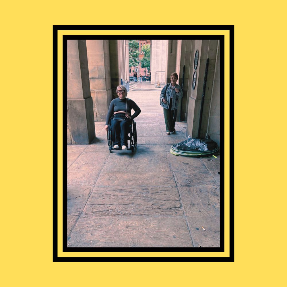 On a yellow background bordered in black, a photograph captures Laura and Melissa beneath a set of pillars. Laura, positioned further back, has close-cropped reddish hair and is focused on taking a picture of Rachel, who is off-screen. In the foreground, Melissa sits more prominently; she has blonde hair, wears black glasses, and is seated in her brown wheelchair, smiling at the camera.