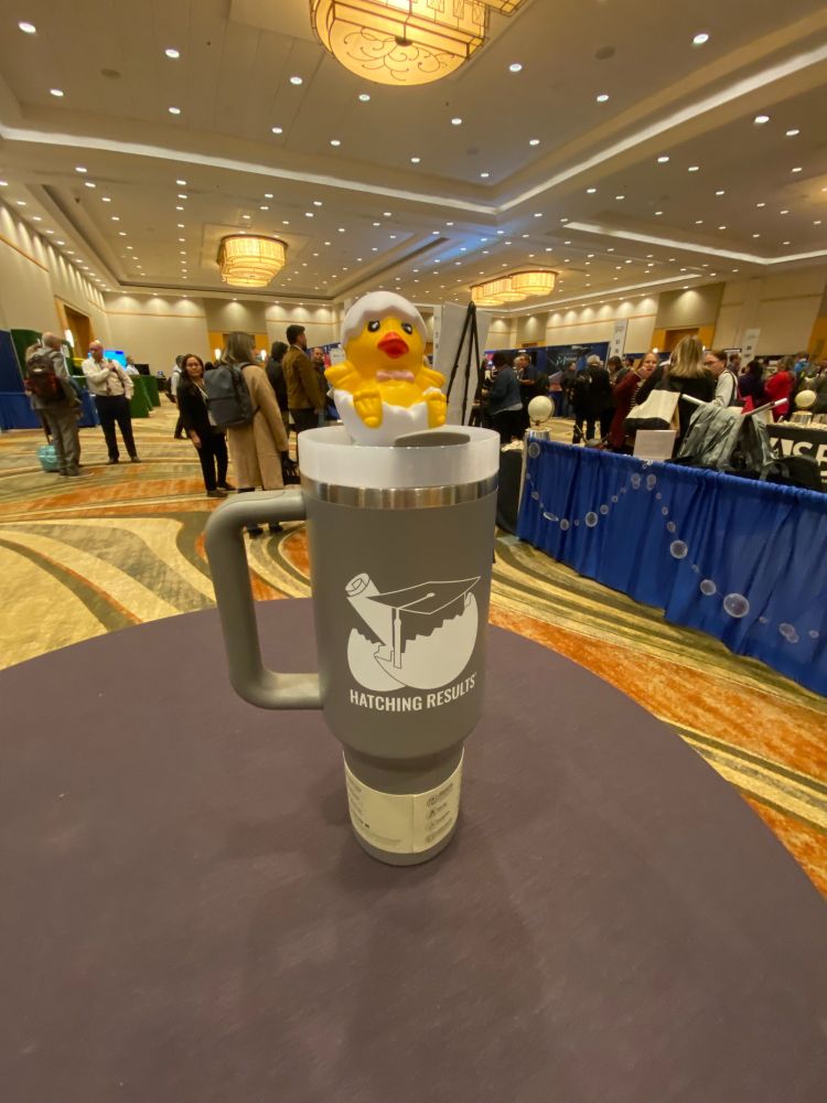 A small yellow chick hatching out of an egg is sitting on a grey Stanley cup with the Hatching Results logo on it. It's on a table with the conference exhibit hall in the background.