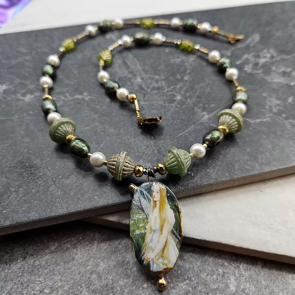 A handmade ceramic focal of an ethereal blonde fairy against a green backdrop, strung on a necklace with green rondelle beads, round white beads, and small gold beads