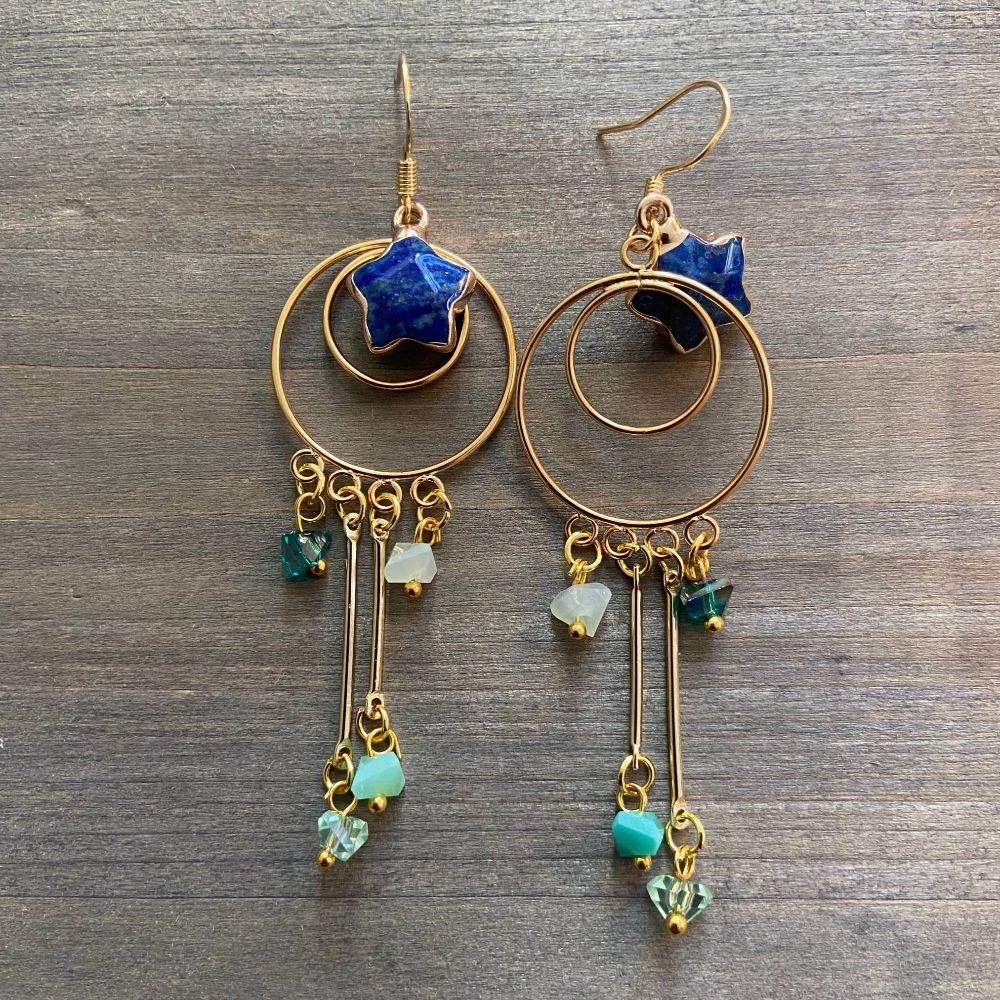 A pair of earrings consisting of concentric golden rings with a lapis lazuli star at the center of the ring, and glass chips in various shades of blue hanging from the rings.