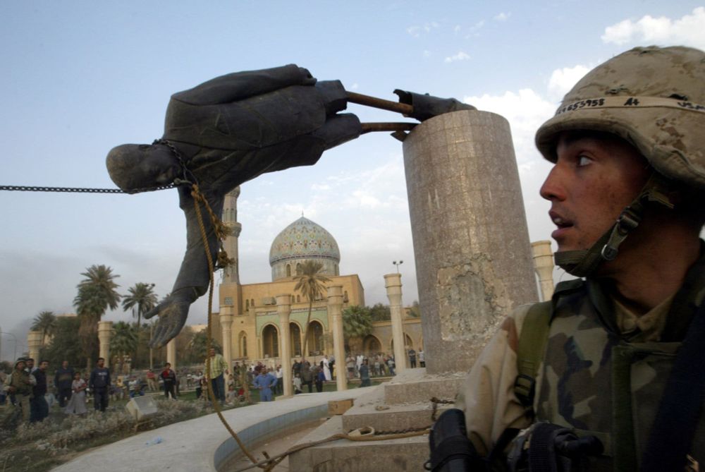 Marines topple a statue of Saddam Hussein on a pedestal in Baghdad, Iraq.  The metal skeleton of the statue is visible, as the exterior metal shell has separated near the knees.  In the back is a domed building and some palm trees.