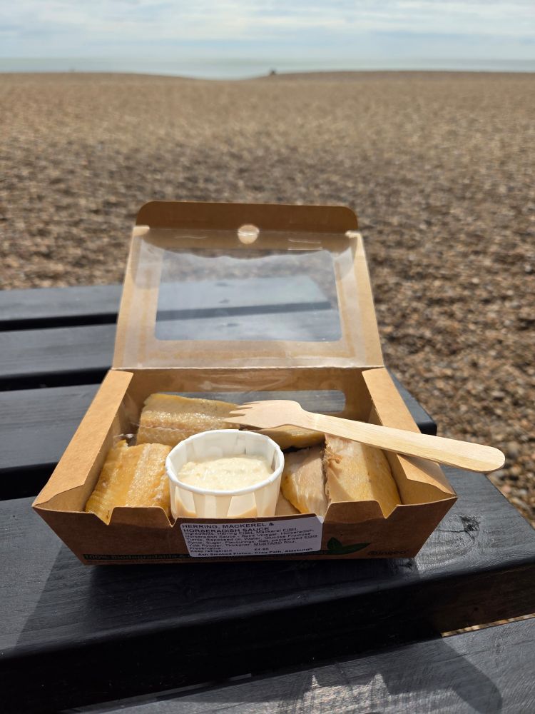 A cardboard box, containing smoked Herring and smoked Mackerel and a pot of horseradish sauce, sits on a black wooden picnic table. Behind it a pebble beach stretches to the sea.