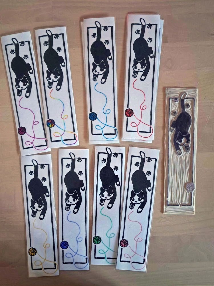A photo of the same bookmarks, but on a table. There's lots of them in different colors. The original lino block is next to them.