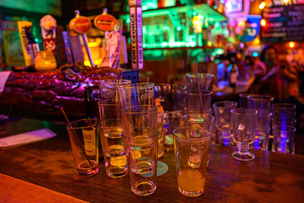 Color photo.  A wooden bar top, upon which are rows and stacks of used pint glasses, with other chalice-style glasses behind.  One of the pint glasses has a small amount of beer left.  Out of focus behind the glasses, on the left, are a row of taps.