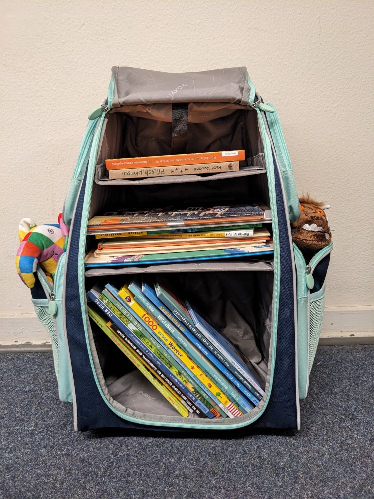 Der blaue Rollkoffer geöffnet: Darin in drei Fächern verschiedene Kinderbücher, teils hochkant stehend, teils quer liegend.