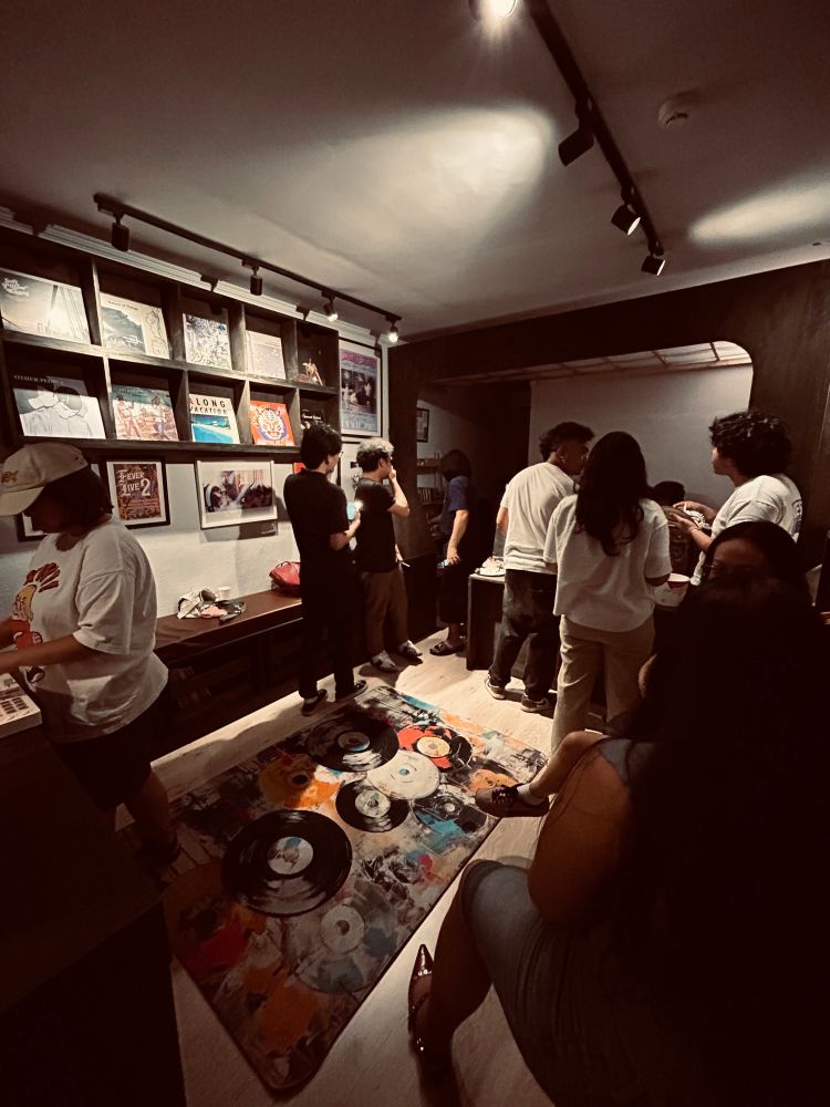 Record store interior. Vinyls on the wall shelf, a carpet with vinyl designs on the floor and people sitting/standing around in the shop