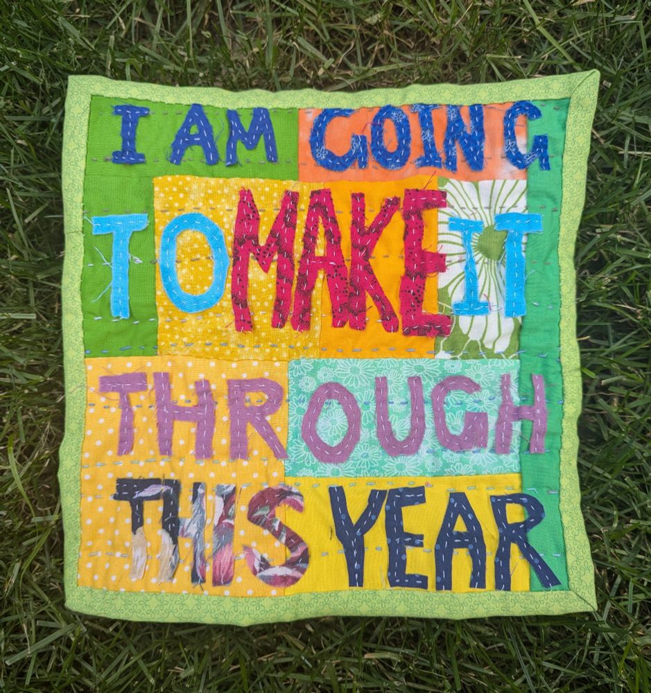 Front of a small hand sewn quilt. Top is green and yellow patchwork with the words "I am going to make it through this year" sewn on in blue, purple and pink fabric.