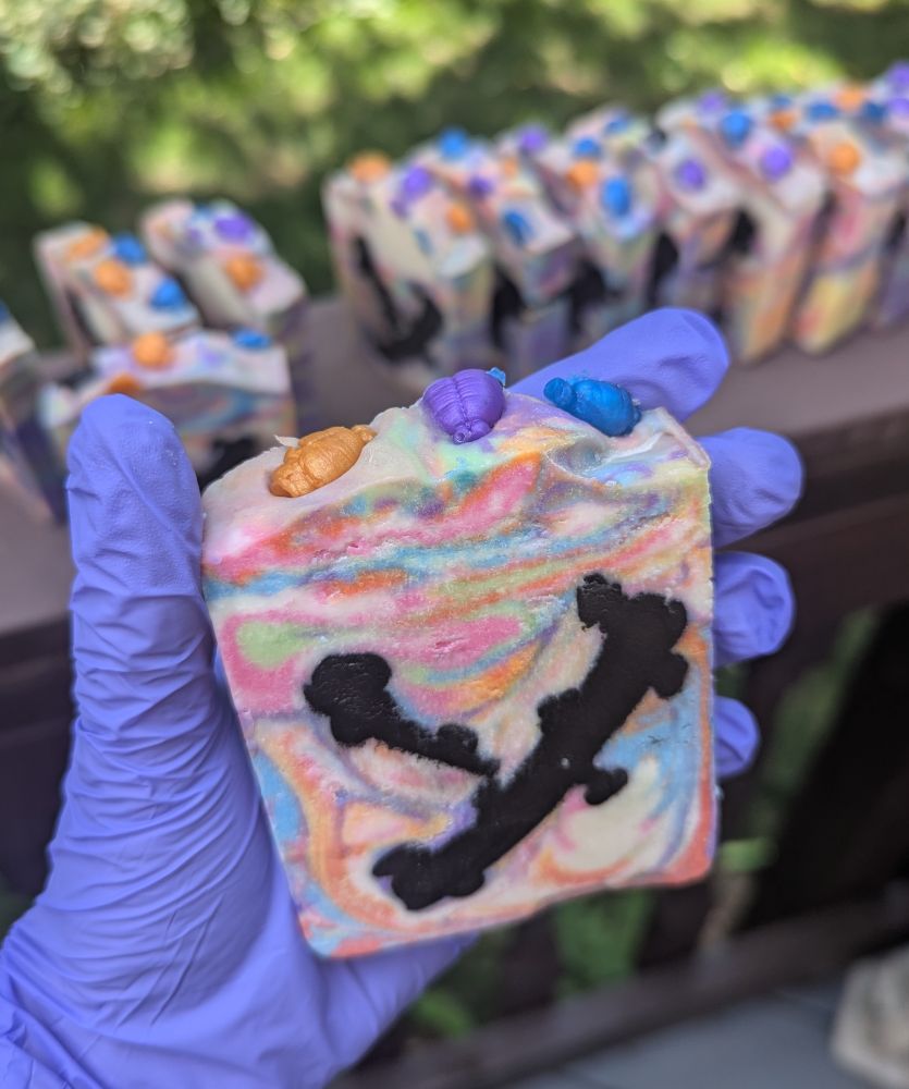 A blue gloved hand holding a rainbow swirl bar of cold process soap.