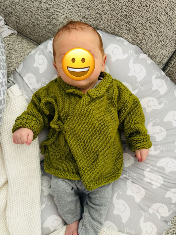 Baby boy wearing a green wraparound sweater that is a little too big still. The sleeves are rolled up a couple times. He’s lying on a grey and white Great Auntie has added a smiley face emoji for privacy. 