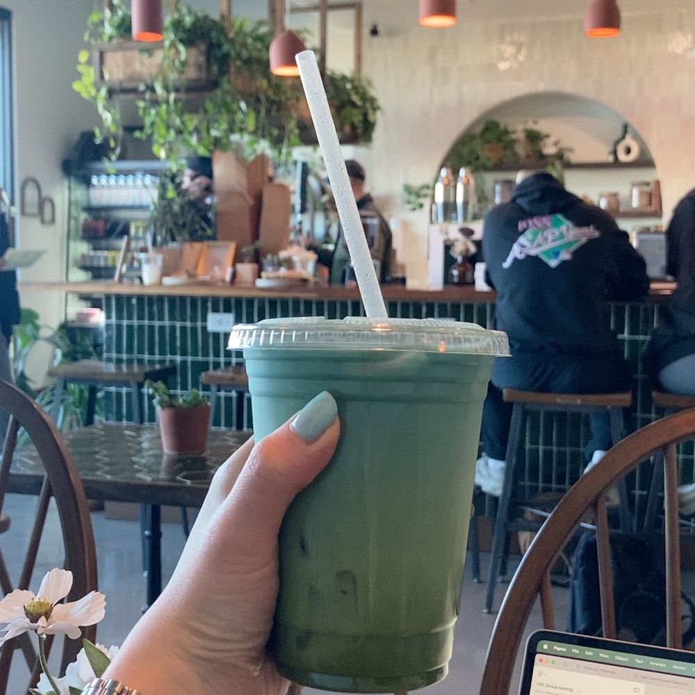 A hand holding a drink that boasts melding shades of aqua to green, held against the backdrop of a coffee shop.