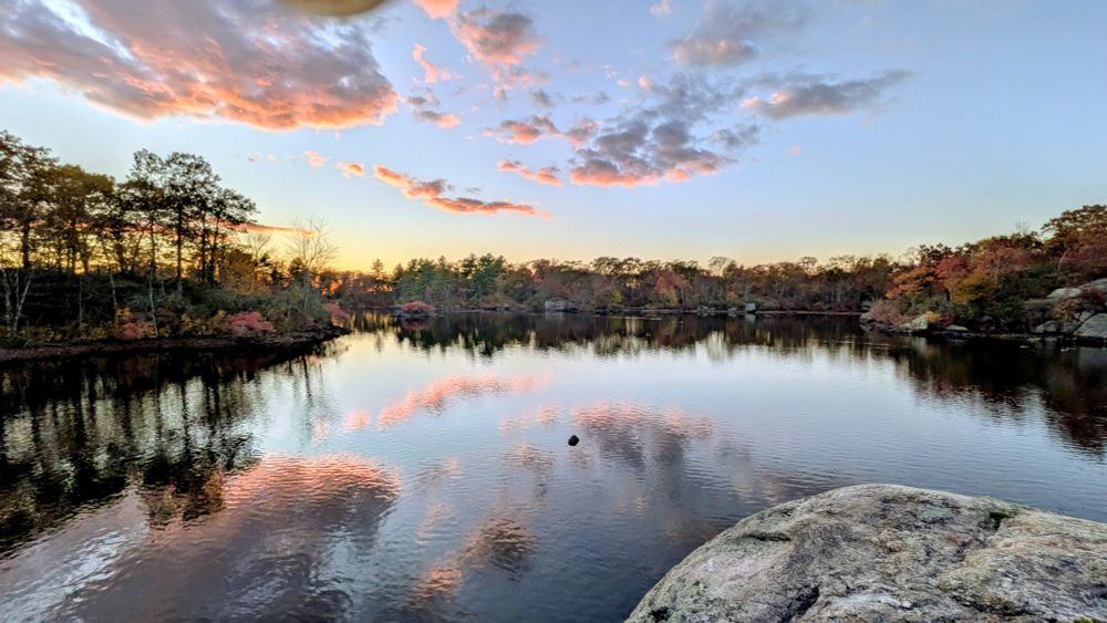 A body of water with the sun setting on the horizon and the clouds reflecting off of the water surface 