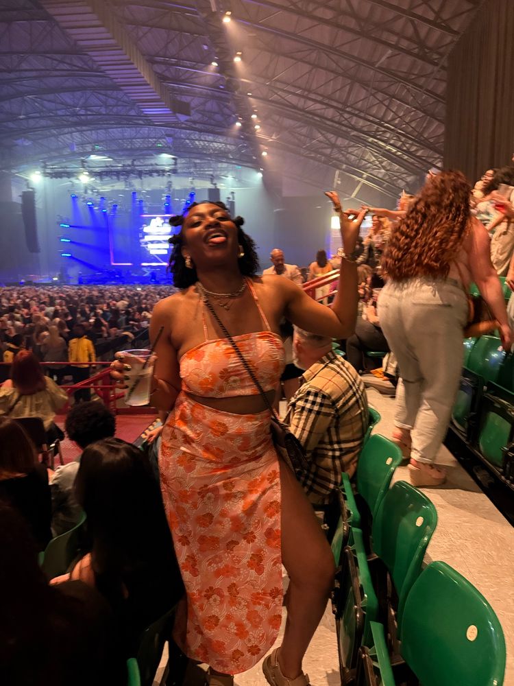 Pearl is wearing a two piece orange pink and white flowery high slit skirt with an alter neck top. She also has a nice sandals and carrying a drink in a cup with a straw. 

She is at a Sean Paul’s concert in Newcastle with her tongue out and deuces sign with her fingers 