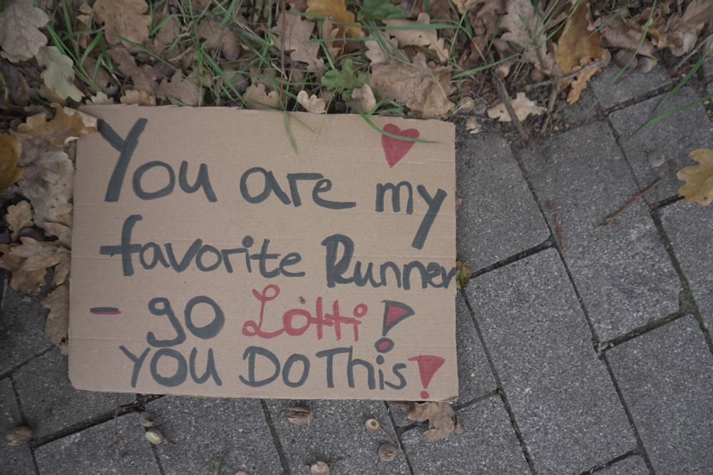 Ein handgeschriebenes Anfeuerungsschild aus Pappe liegt auf einem Gehweg neben Herbstlaub. Darauf steht der englische Text: "You are my favorite Runner – go Lotti! YOU DO This!". Der Name "Lotti" ist in Rot geschrieben und über dem Text ist ein kleines rotes Herz gemalt.