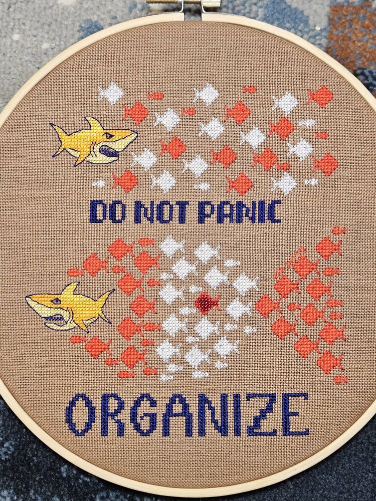 Photo of an original cross stitch design in a hoop.  The fabric is a 28 count warm milk mocha colour.  The top half of the design features an orange shark chasing a pool of random red and white fish, with the words underneath "DO NOT PANIC"
The lower half of the design depicts the orange shark fleeing from the organized red and white fish that have come together in the shape of a larger fish.  Moreover, the organized shape is in the colours of the Canadian Flag, with a sparkly red beaded fish in the centre of the white field where the Canadian Maple leaf usually is.  At the base is the large stitched word "ORGANIZE".
It's a reminder that as individuals sometimes tyranny seems insurmountable,  but when we band together, we are strong and will defeat the orange menace.