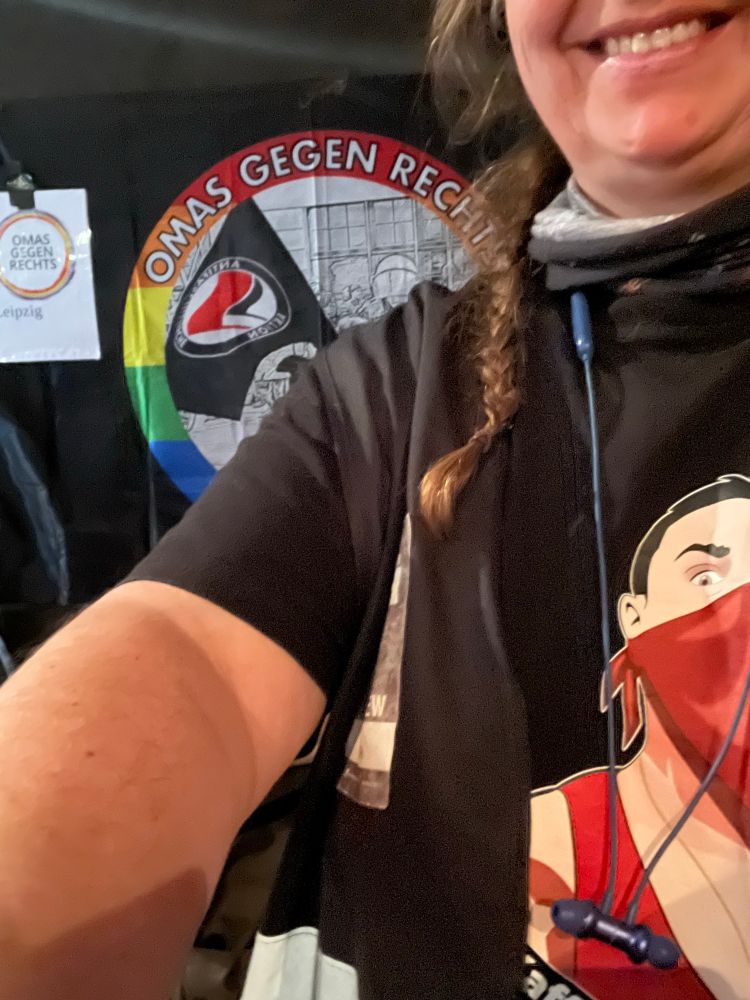 Das Foto zeigt ein Selfie von mir, ich lächele und bin nur teilweise im Bild zu sehen ist. Ich trage ein schwarzes T-Shirt mit einer gezeichneten Figur, die ein rotes Oberteil und ein rotes Tuch als Gesichtsmaske trägt. „Kein Kaffee  für Nazis’Um den Hals hängen blaue In-Ear-Kopfhörer.

Im Hintergrund hängt ein großes Banner mit der Aufschrift „OMAS GEGEN RECHTS“, dazu ein Regenbogen-Hintergrund und ein Antifa-Symbol. Links daneben ist ein kleineres Schild mit derselben Aufschrift, ergänzt um den Ortsnamen „Leipzig“.

👉 Insgesamt vermittelt das Bild eine klare antifaschistische Haltung und zeigt die Person in einem solidarischen Umfeld, das sich gegen Rechts engagiert.