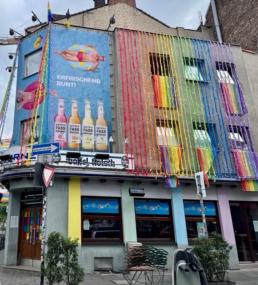 Blick auf ein Stadthaus. Unten ist Gastronomie. Ab der ersten Etage hängt aus fast jedem Fenster die Regenbogenfahne und gleichzeitig hängen von der Dachrinne regenbogenbunte Stoffstreifen.