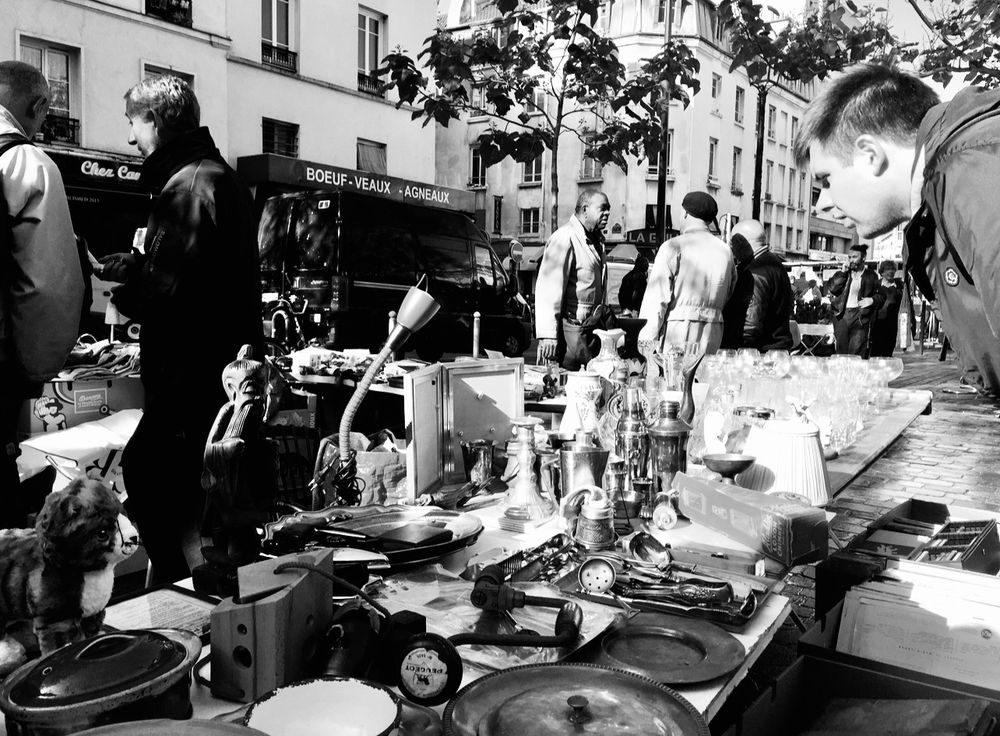For Urban Gaze, Humans of Bluesky and Classic Mono, a black-and-white photograph, wide view of an antique stall on a street in Paris. A long table is filled with antiques and collectibles, including, what looks like a tempting, silly, stuffed cat. On the left two men are walking past, in the centre three gentlemen are having a conversation and on the right, the head of a man looking intently something of interest. 
