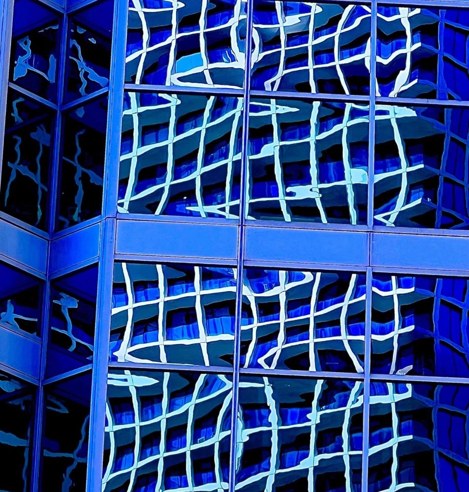 For the Blue Sky Art Show theme Lines, a colour photograph zoom view taken in downtown Toronto, of the side of a royal blue glass, high-rise office tower in downtown Toronto.   The distorted, square white window frames of the building across the way are reflected in the glass, creating an interesting wavy line abstract effect. 