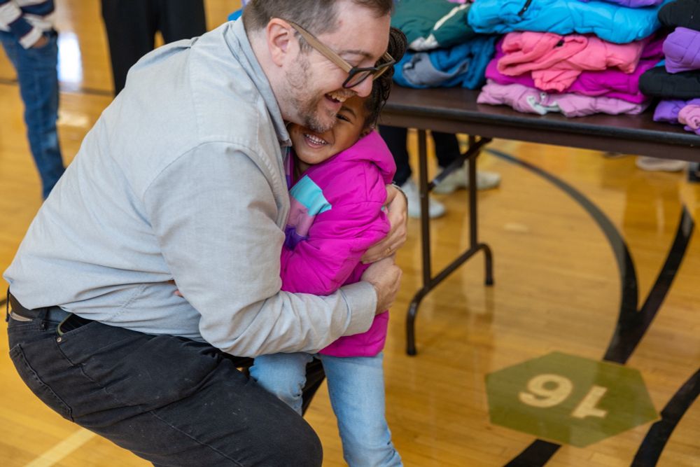 An adult (me) crouches down and hugs a young child in a bright pink winter coat inside a gymnasium. Both are smiling joyfully. Behind them is a table stacked with colorful winter coats, and the gym floor markings are visible beneath them.