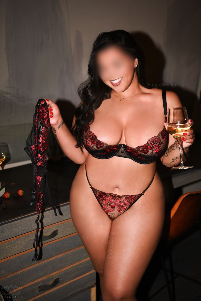 Paige Savage smiling in red and black embroidered lingerie, holding a matching garter belt in one hand and a glass of white wine in the other, standing by a desk with scattered strawberries in a softly lit NYC suite.