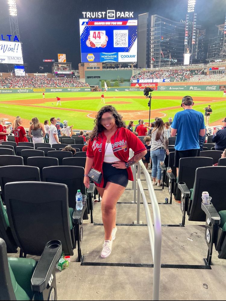 A woman poses at Truist Park during a Braves night game, standing on stadium steps with the baseball field lit behind her. She wears a red Braves jersey over a white top and black shorts, paired with white sneakers and a crossbody bag. Her smile is bright, radiating playful, sporty charm.