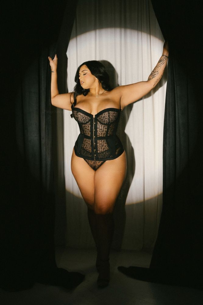 A confident woman posing in a black lace corset and matching panty, standing between dark curtains. She holds the curtains open with both hands, creating a spotlight effect on her hourglass figure. The setting is elegant and dramatic, emphasizing self-assurance and empowerment. 