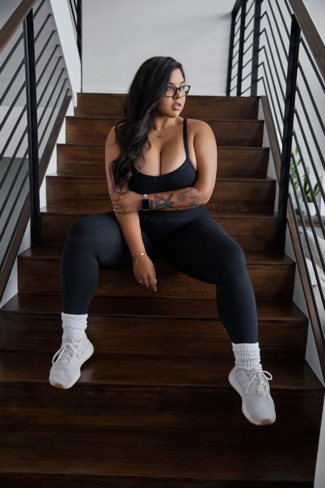 Paige Savage, a woman with long, dark wavy hair and glasses is sitting on a modern wooden staircase with metal railings. She is wearing a black fitted workout outfit, white socks, and white sneakers. Her arms are crossed over her body with one hand resting on her opposite arm, and she has a confident, relaxed expression as she looks off to the side. She has accessorized with a smartwatch and bracelet. The background is minimalistic indoor space with neutral tones and sleek design elements. 
