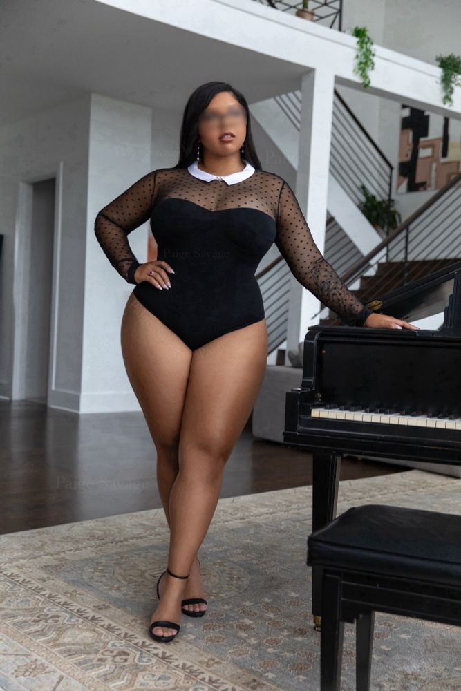 A curvy woman stands beside a grand piano in a high-ceilinged modern loft, wearing a sheer black bodysuit with a white collar and heels. Her eyes are softly blurred, adding to the sense of discretion. She exudes quiet luxury and is poised, magnetic, and completely at ease in her power.