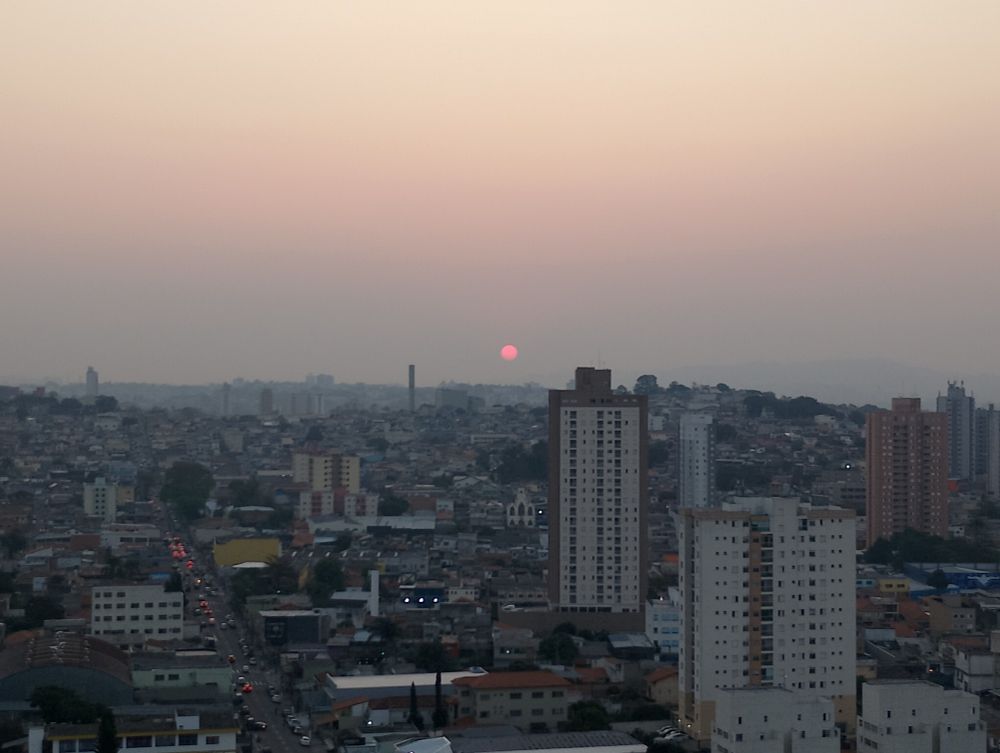 Vista da sacada do meu apartamento, com vários prédios, ruas e casas. Ao fundo, o céu está poluído, com uma tonalidade cinza no horizonte que vai clareando para o rosa e o bege à medida que o olhar sobe. Próximo ao horizonte, no meio da imagem, está o Sol, visto como um círculo vermelho-claro no fundo cinza.