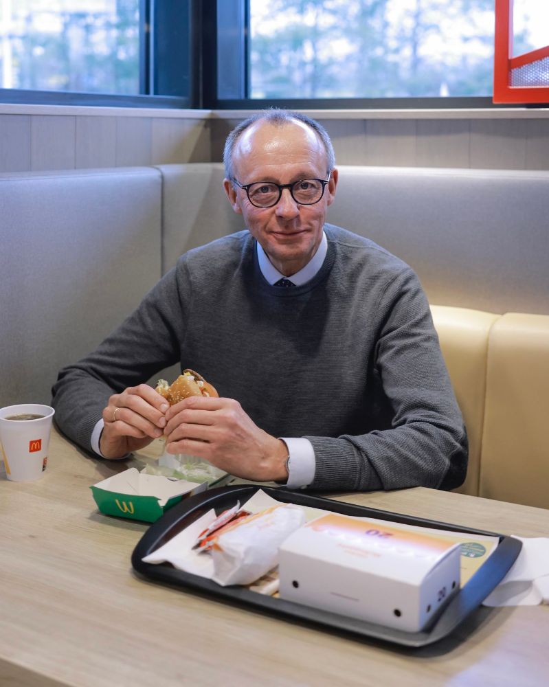 Friedrich Merz sitzt offenbar in einem McDonalds-Restaurant und hat einen Burger in der Hand. Darunter die offene Burger-Packung mit einem gut sichtbaren Logo. Daneben eine Getränkebecker ebenfalls mit Logo.