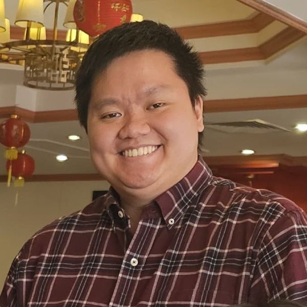 Chrispy, an Asian man with black hair smiles at the camera. He is wearing a red checkered plaid shirt. 