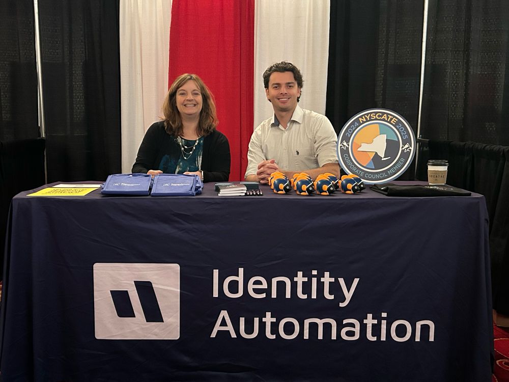 Susan Bearden at Jack Welling sit at the Identity Automation booth behind a table with  purple tablecloth. There is swag promoting IA services including comprehensive IAM, cybersecurity, and PHISHID anti-phishing protection.