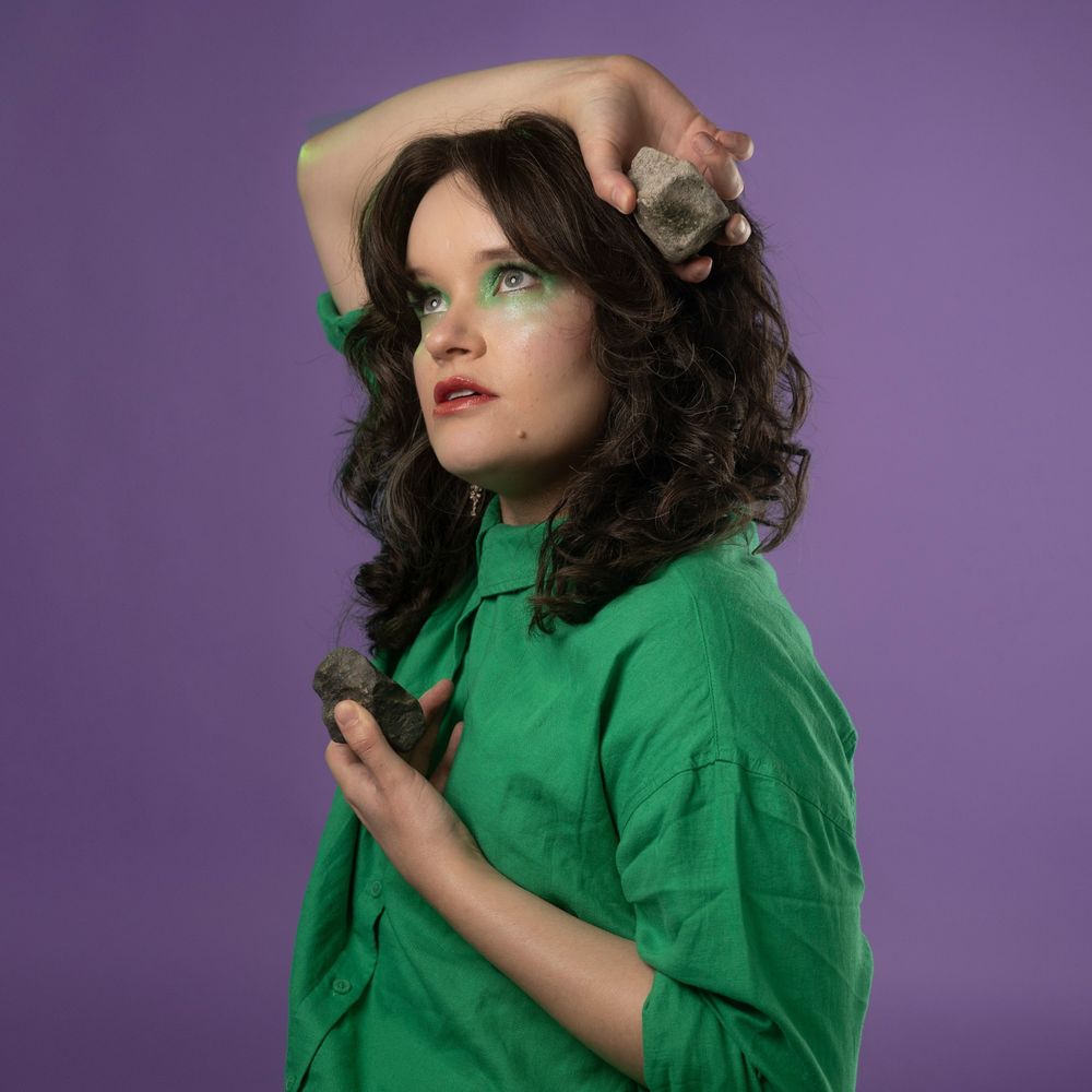 A women in a green shirt with green eye shadow. She is looking up, posing, while holding two rocks in her hands. She is photographed against a lilac background 