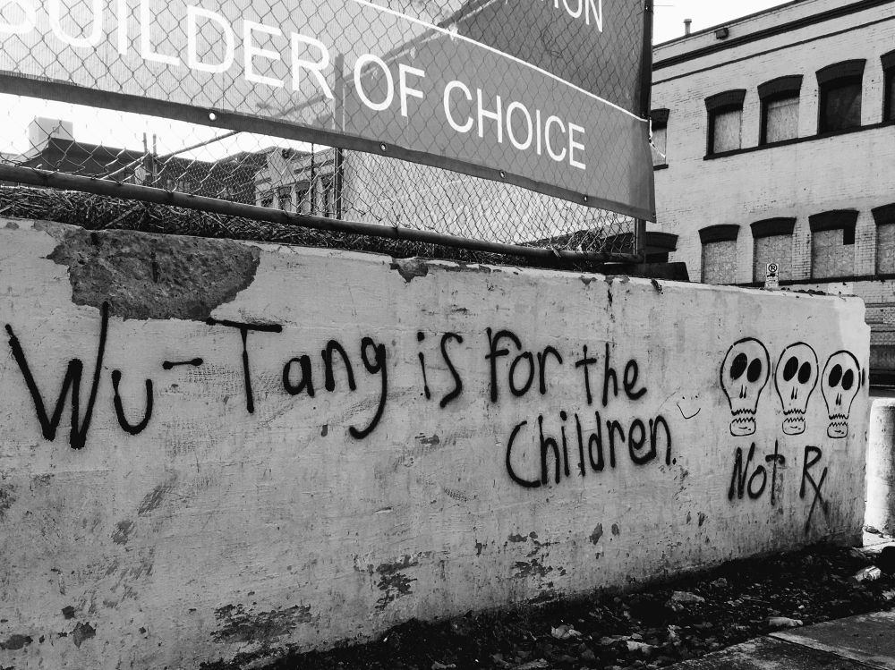 Black and white photo of a concrete wall with spray painted “Wu Tang is for the children”. A banner above it on a new fence says “builder of choice”. An older building with boarded up windows provides a backdrop. 
