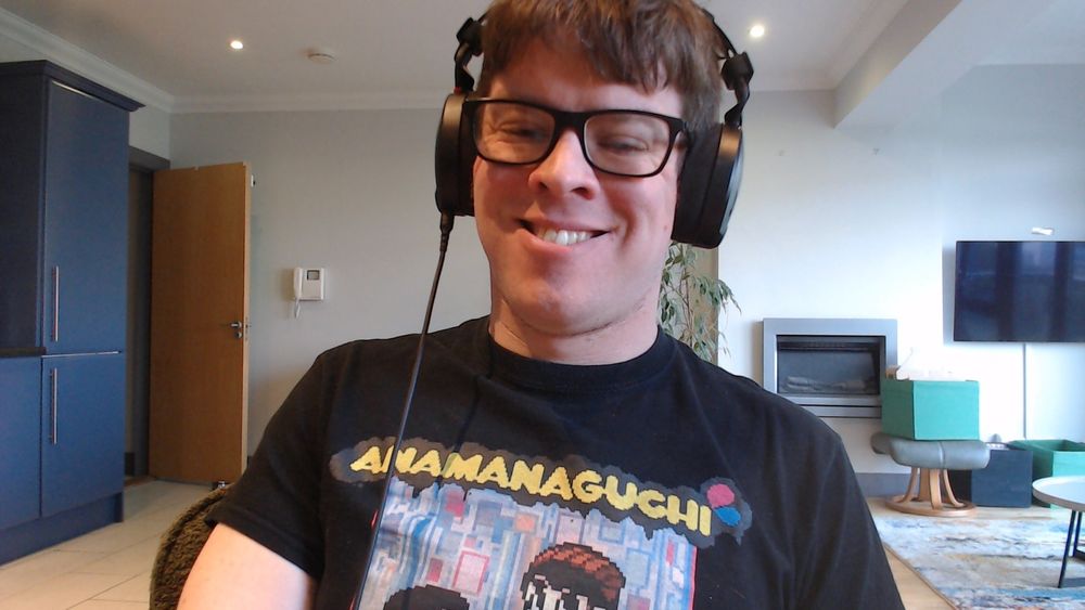 Person with brown hair and glasses, smiling, wearing a pair of black headphones and an Anamanaguchi t-shirt.