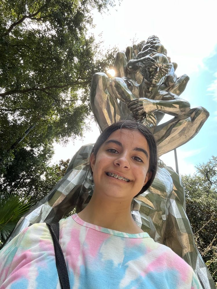 A smiling adolescent girl in tie dye shirt  photographed in front of a silver sculpture of many iterations of a crouched figure at such an an angle that it appears the crouched figure is emanating from her head