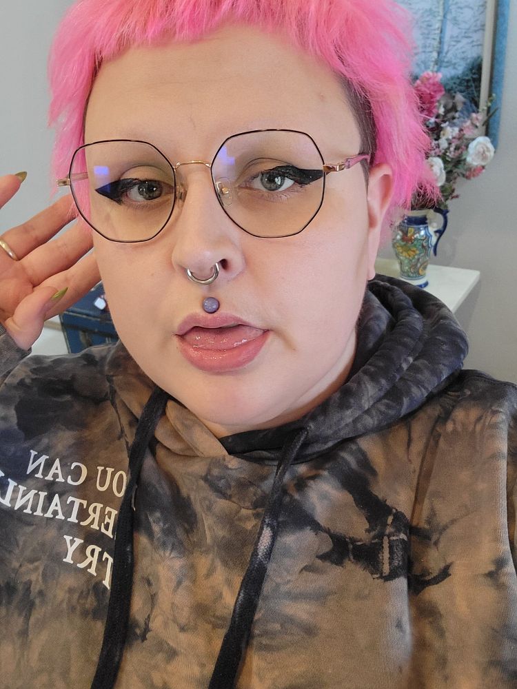 A white agender person is looking at the camera with their tongue partially sticking out. They have pink hair, are wearing glasses and are a tie dye critical role hoodie that says "you can certainly try"