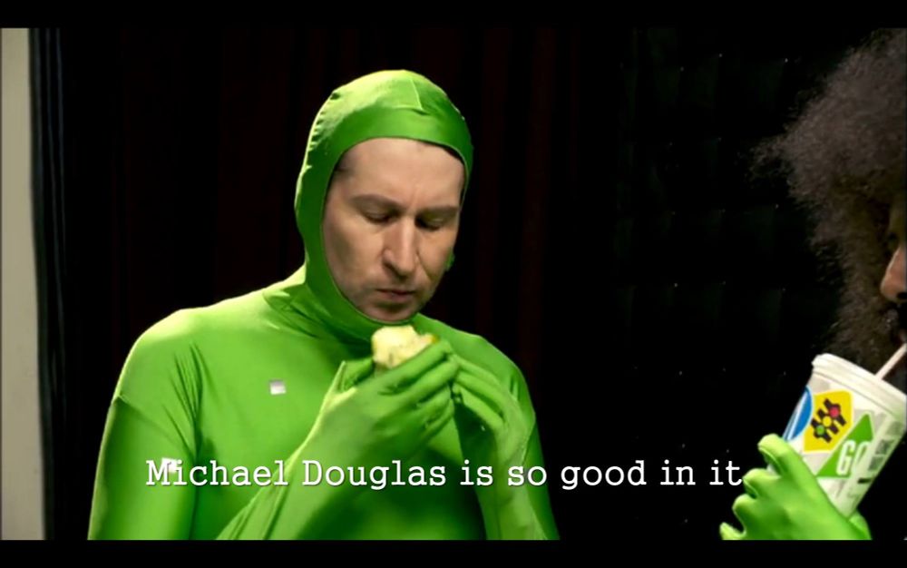 Scott Aukerman in an all green jumpsuit saying Michael Douglas is so good in it. 
