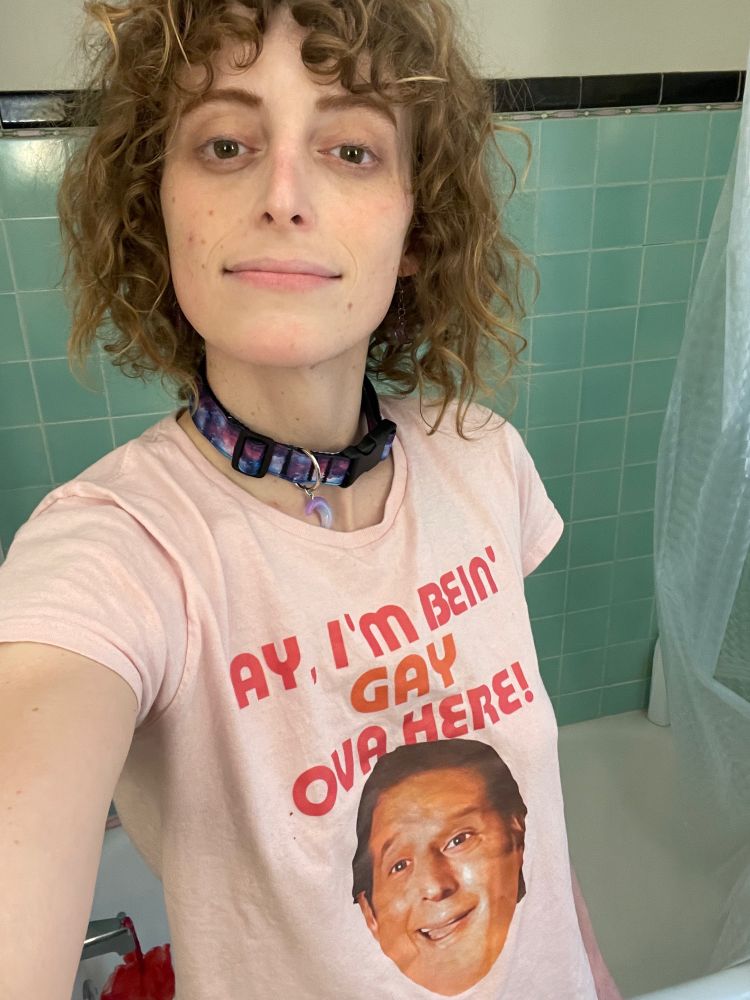 wearing a pink shirt that reads ‘ay, im bein gay ova here’ and wearing a purple collar with a moon dangling from the end.