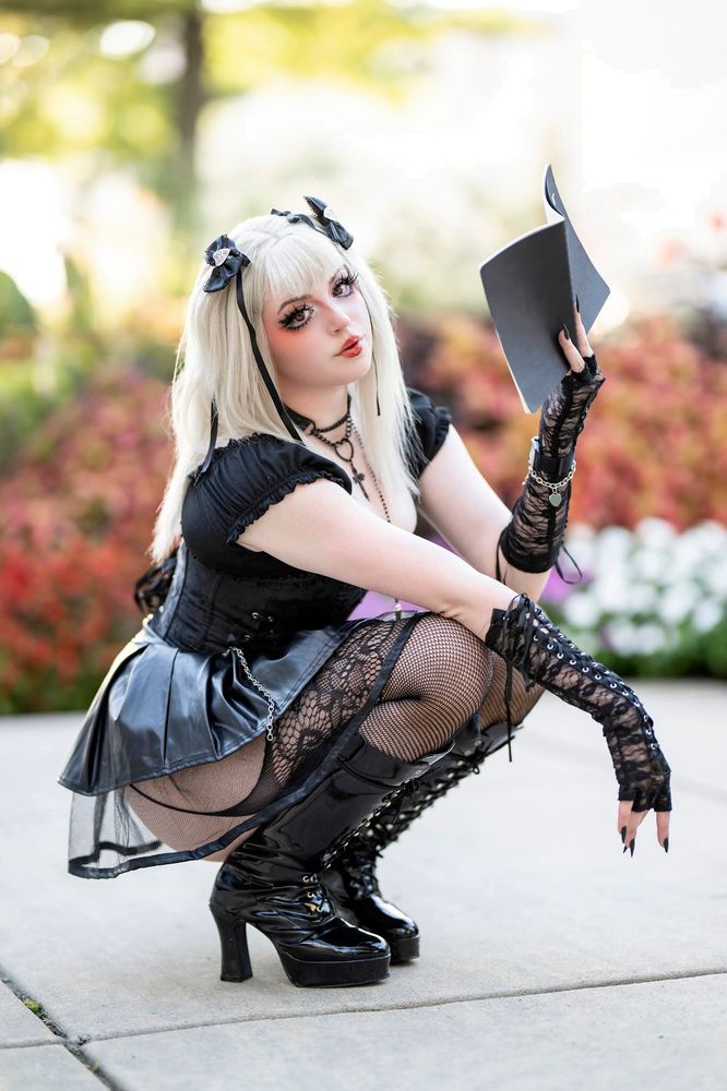 Valentine is in a squat position, cosplaying Misa Amane from death note. The entire outfit is black in color consisting of a corset, cross choker, lace arm socks, gogo boots, fishnet leggings, blonde 22in wig, two black hair ribbons and holding a death note in their left hand. The background is lined with flower bushes. 