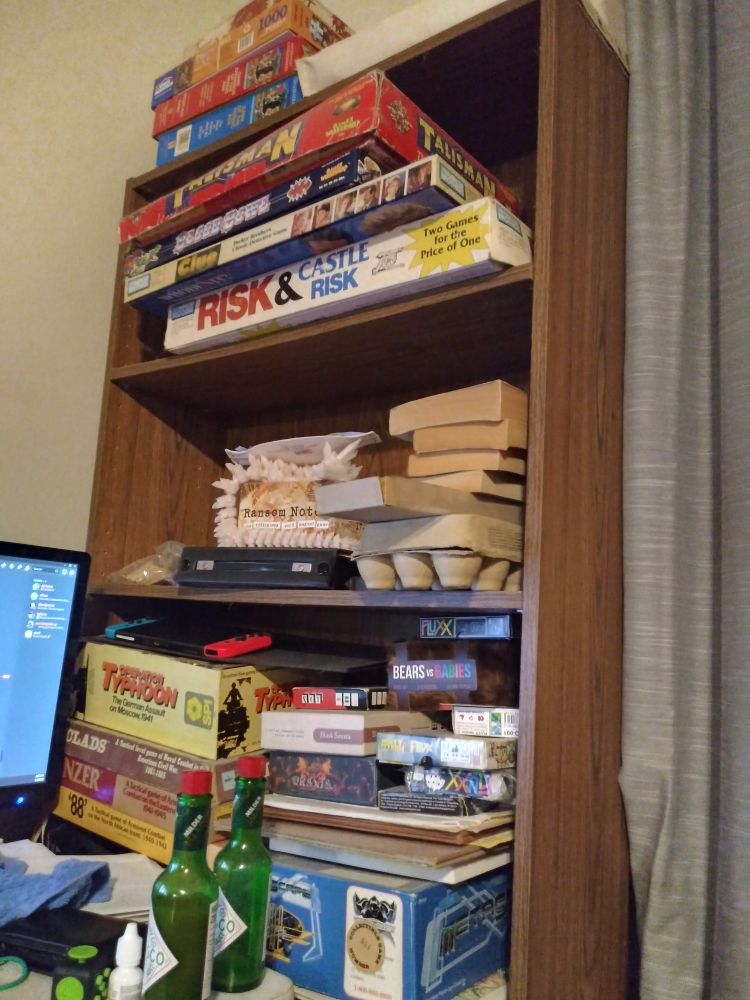 A picture of a set of shelves haphazardly filled with board games, card games, books, a Nintendo Switch, one of those articulated 3d printed dragon things, and other miscellaneous objects. Also visible are hot sauce bottles, a fidget cube, part of a computer screen, a hair tie, and other random things on the corner of a desk.