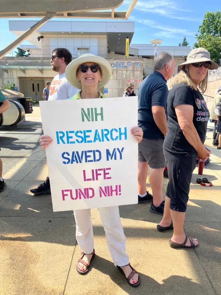 A person attending the NIH vigil holds a sign saying "NIH research saved my life/Fund NIH!"