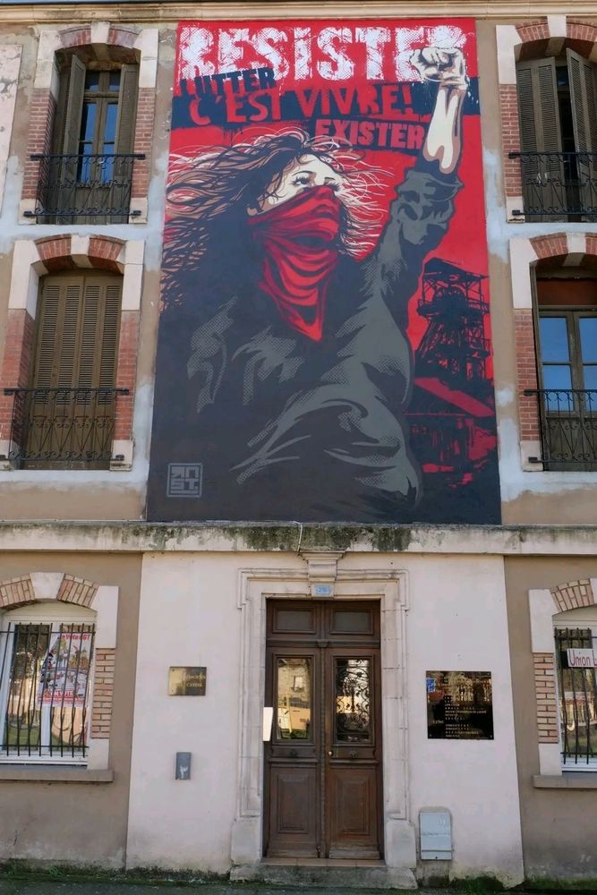 photo d'un immeuble avec un tableau géant entreposé dessus. on y voit une personne levant le poing gauche avec un foulard sur le bouche. au-dessus, on lit : "Résister, lutter, c'est vivre ! Exister".
