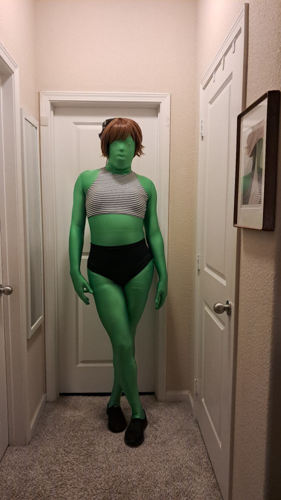 A guy in a zentai and shirt brown win poses in a black-and-white striped bikini top and black bottoms.