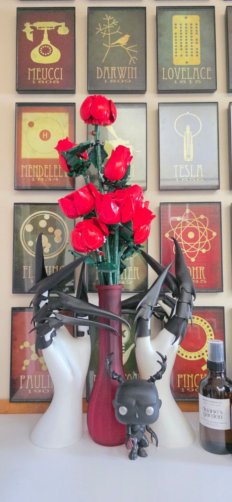Photo of a frosted red vase full of bright red Lego toy roses. At the base of the vase are two white jewelry display hands wearing long black costume armor claws, as well as a funko pop of a Wendigo from the TV series 