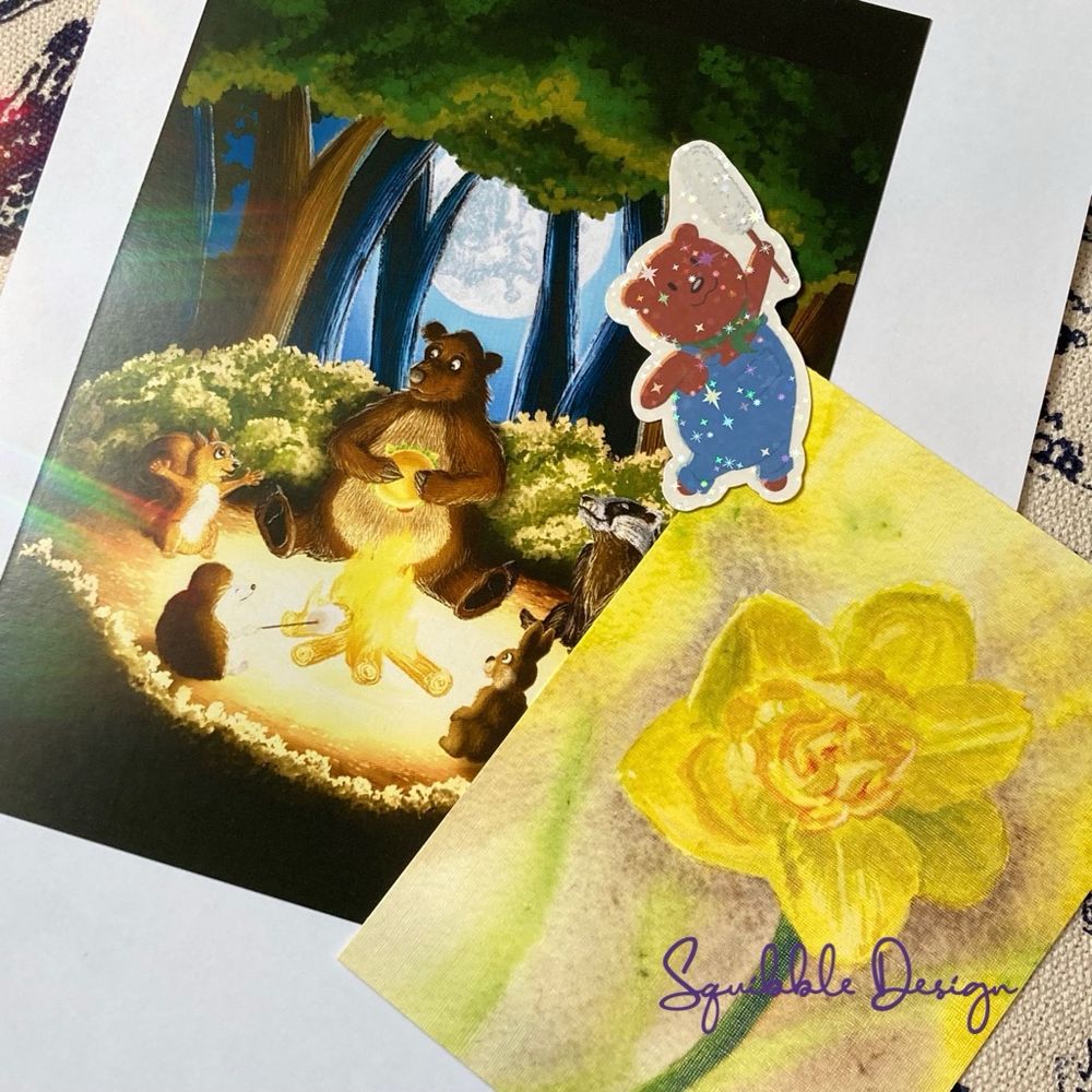Art print of animals round a camp fire, a sticker of a bear in overalls, and a postcard with a painting of a daffodil.