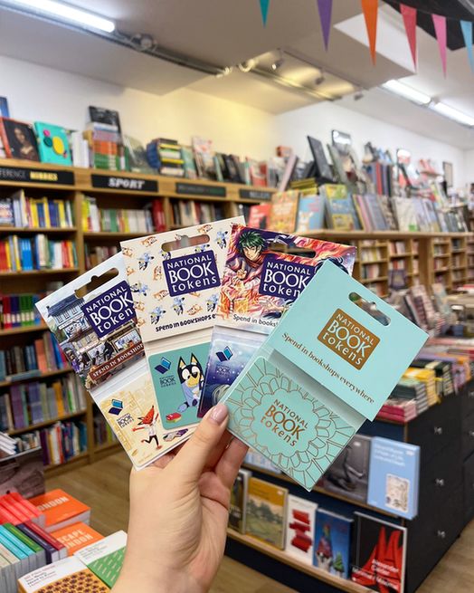Four new National Book Tokens gift card designs featured in a busy bookshop with colourful displays and bunting.