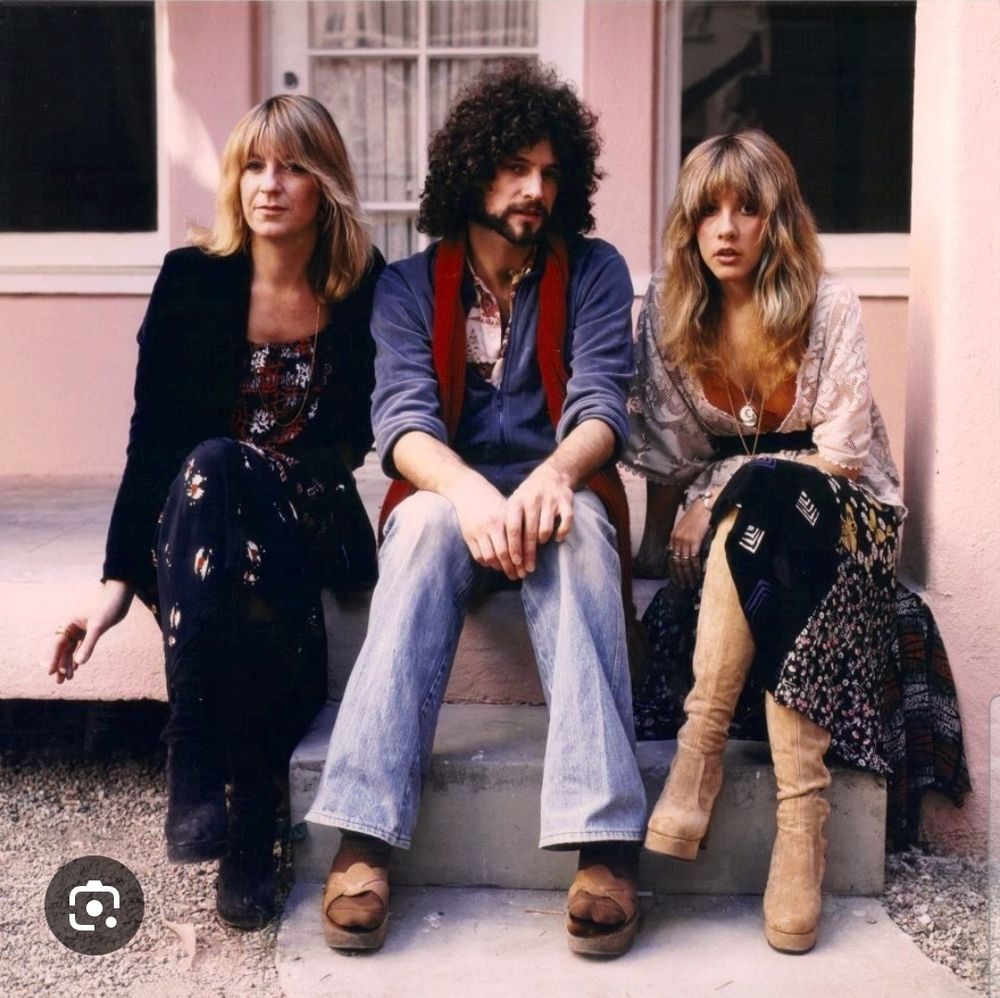 Christine McVie, Lindsey Buckingham, and Stevie Nicks of Fleetwood Mac, sitting on a doorstep of a pink building. 